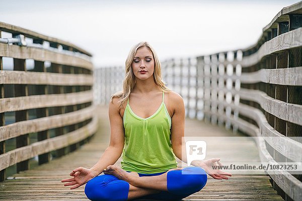 Portrait einer jungen Frau  die Lotus-Yoga-Pose auf einem hölzernen Pier praktiziert.