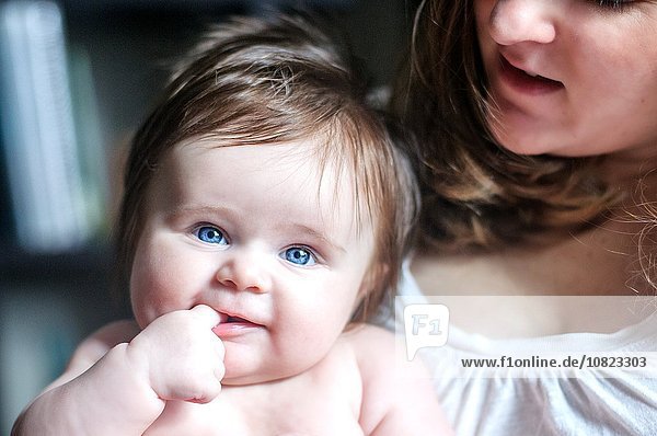 Mutter hält Baby Tochter mit Finger im Mund und schaut lächelnd in die Kamera.