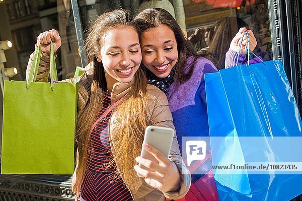 Young female adult twins in city taking smartphone selfie with shopping bags
