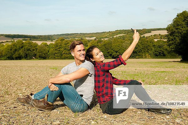 Junges Wanderpaar sitzt Rücken an Rücken im Feld und nimmt Smartphone Selfie  Great Missenden  Buckinghamshire  U.K.