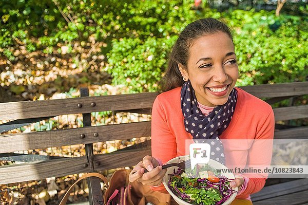 Young woman sitting on park bench eating lunch