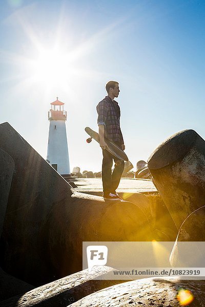 Junger Mann mit Skateboard  stehend auf dem Wellenbrecher beim Leuchtturm