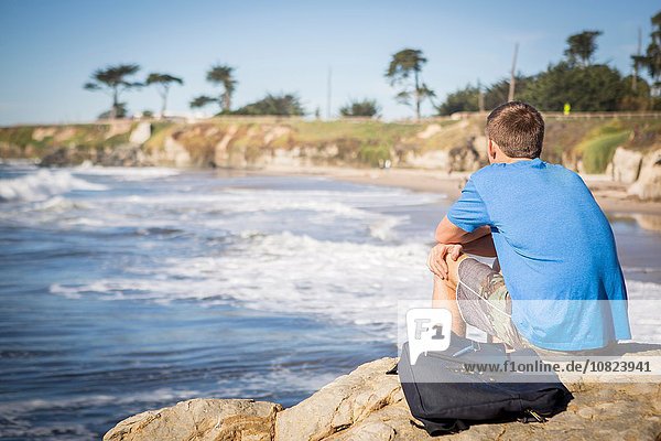 Young man sitting by rock  looking out to sea  rear view