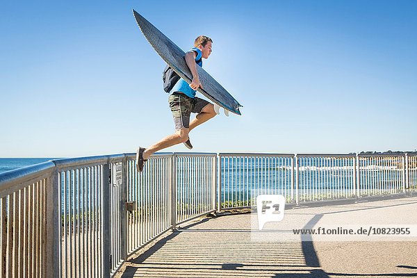 Junger Mann  trägt Surfbrett  springt vom Geländer neben dem Meer