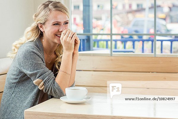 Junge Frau am Tisch sitzend mit Tee und Untertasse  auf Ellenbogen liegend  lächelnd wegschauend