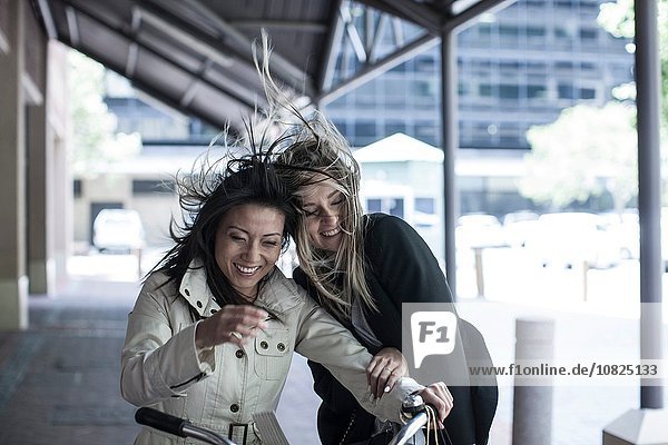 Two women friends with flyaway hair laughing on bicycle in city