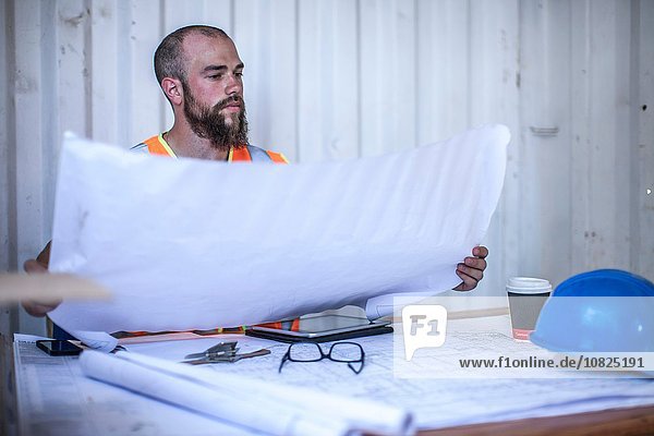 Construction worker sitting at desk looking at blueprint in portable cabin