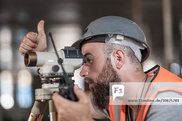 Young male surveyor looking through theodolite giving thumbs up on construction site