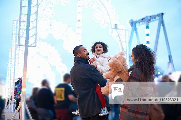 Mother and father in amusement park carrying smiling girl and teddy bear