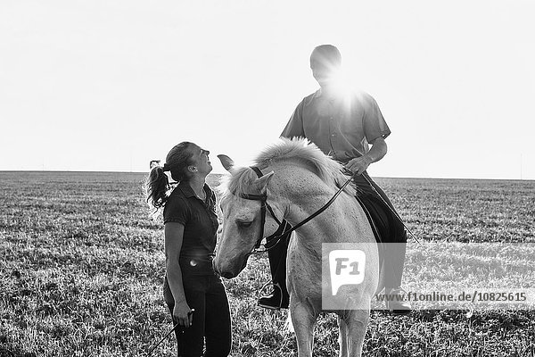 Schwarzweißbild einer Frau  die mit einem Mann auf einem grauen Pferd im Feld plaudert.