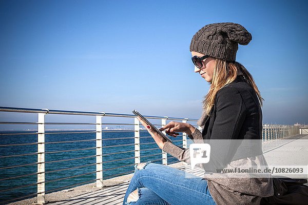 Seitenansicht der jungen Frau am Meer sitzend mit Hilfe eines digitalen Tabletts