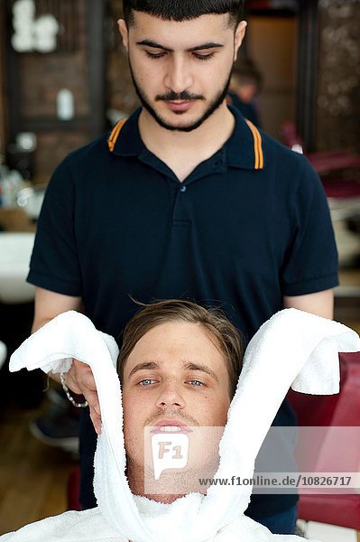 Barbier wickelt das Gesicht eines jungen Mannes in ein frisches Handtuch und schaut nach unten.