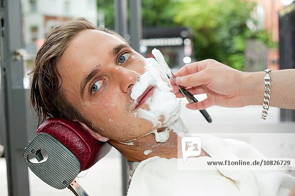 Junger Mann im Friseurladen  das Gesicht mit Rasiercreme bedeckt  die mit einem Rasiermesser weggeschaut wird.