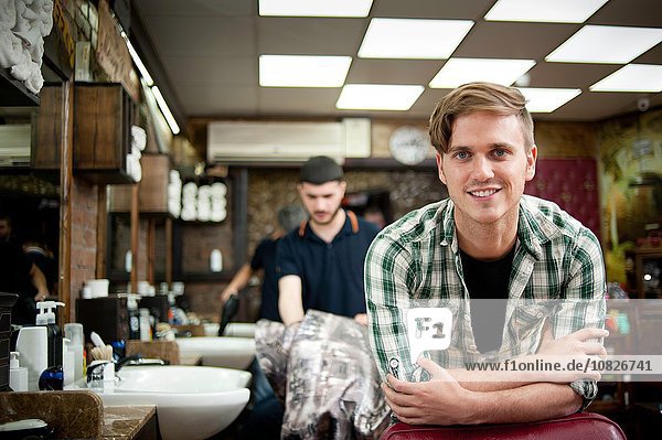 Junger Mann im Friseurladen ruht auf Ellbogen und schaut lächelnd in die Kamera.