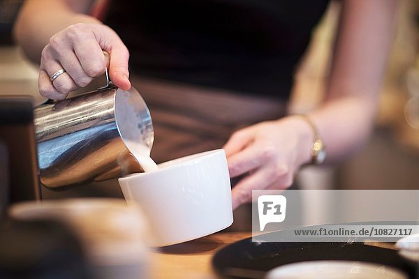 Abgeschnittener Schuss einer Kellnerin  die Milch in eine Kaffeetasse im Café gießt.