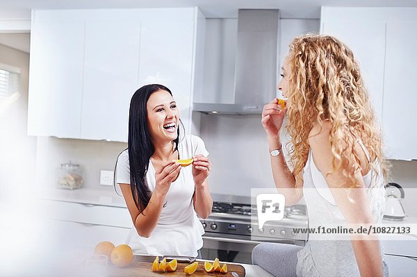 Two young female friends eating breakfast oranges at kitchen counter