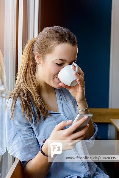 Junge Frau hält Smartphone und trinkt Kaffee