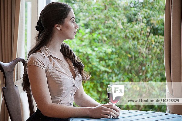 Junge Frau sitzt am Tisch vor dem Fenster und hält ein Glas Wein.