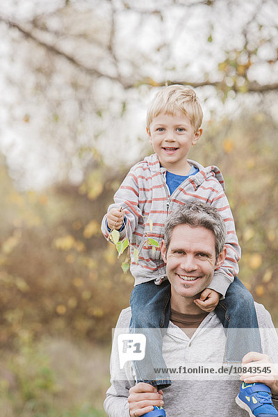 Portrait lächelnder Vater mit Kleinkind-Sohn auf den Schultern im Herbstpark