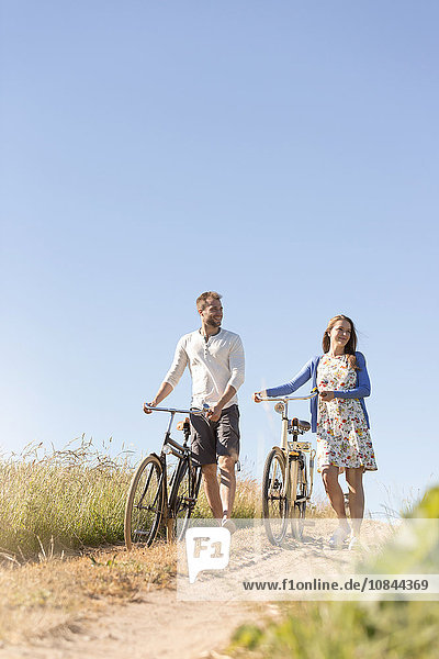 Junges Paar geht mit dem Fahrrad auf dem Feldweg unter dem sonnigen blauen Himmel spazieren