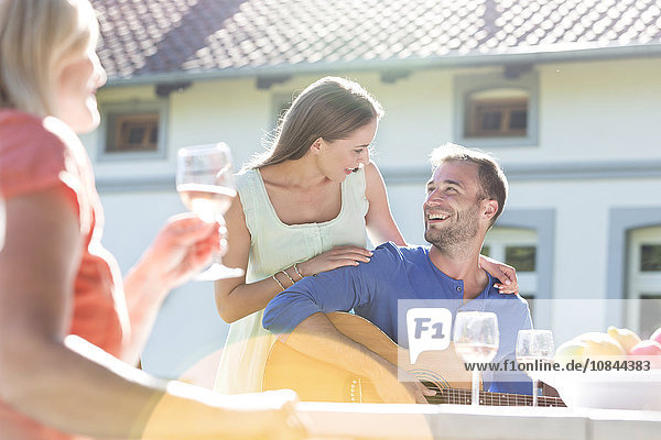 Lächelndes junges Paar spielt Gitarre und trinkt Wein auf der Sonnenterrasse.