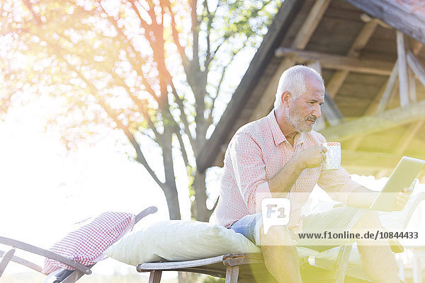 Senior Mann trinkt Kaffee und benutzt Tablet PCn auf einem Sessel im Garten.