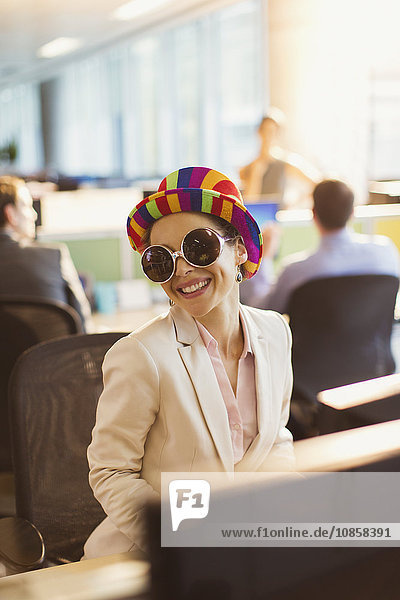 Portrait of smiling businesswoman in silly sunglasses and striped hat working in office