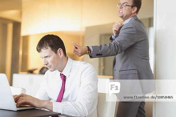 Angry businessman aiming rubber band at unsuspecting businessman working at laptop