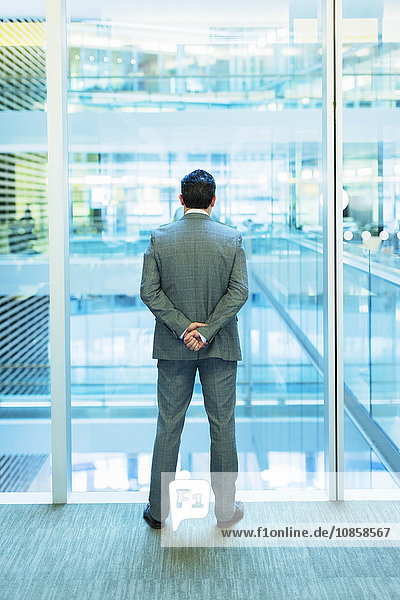 Businessman standing at atrium window with hands behind back