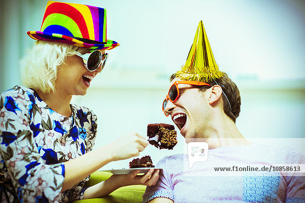 Verspieltes Paar mit Partyhüten und Sonnenbrille beim Schokoladenkuchenessen