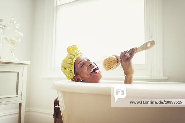 Verspielte reife Frau singt in die Loofah-Bürste in der Badewanne.