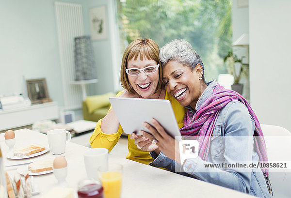 Lachende reife Frauen teilen sich Tablet PCn am Frühstückstisch
