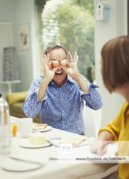 Playful mature man covering eyes with eggs at breakfast table