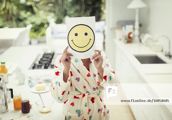 Portrait of women holding smiley face printout over face in kitchen