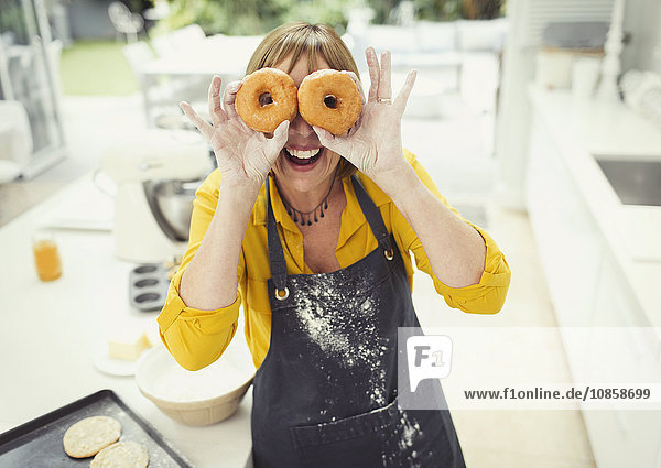 Portrait playful mature woman covering eyes with donuts in kitchen