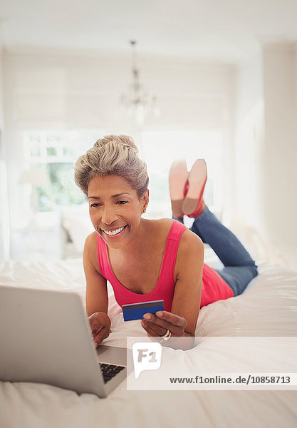 Reife Frau mit Kreditkarte online einkaufen mit Laptop im Bett