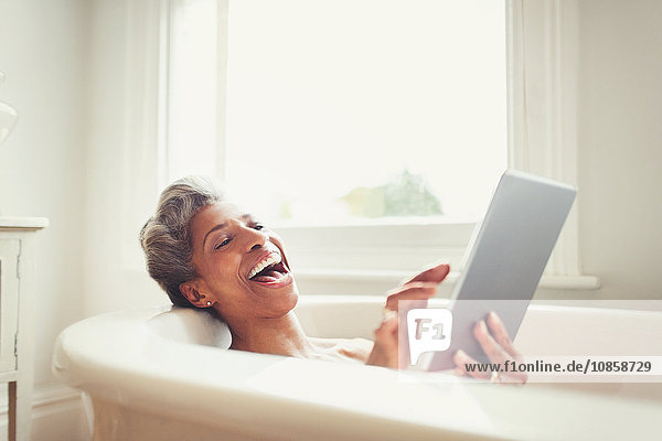 Lachende reife Frau mit digitalem Tablett in der Badewanne