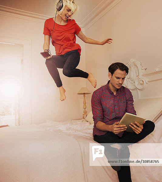 Playful wife jumping on bed listening to music behind husband using digital tablet