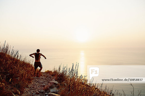 Male runner with backpack descending craggy trail overlooking sunset ocean