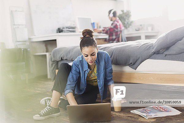 Female college student with coffee using laptop on bedroom floor