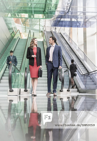 Corporate businessman and businesswoman talking below escalators in modern lobby