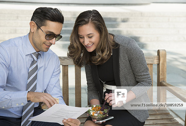 Corporate businessman and businesswoman eating lunch and reviewing paperwork on bench