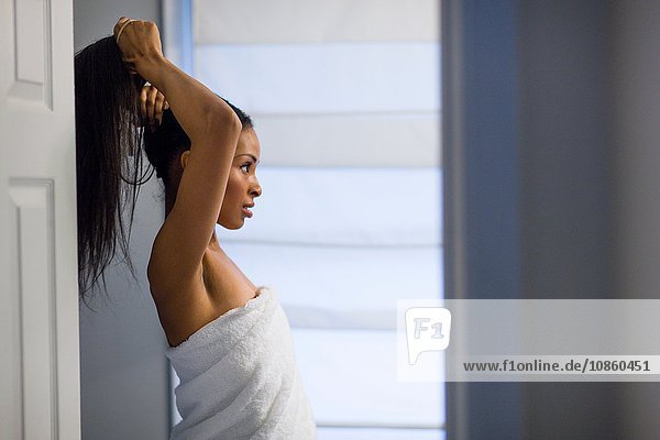 Junge Frau in weißes Handtuch gehüllt  das lange schwarze Haare im Badezimmer zusammenbindet