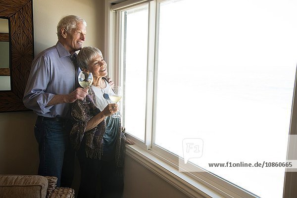 Älteres Ehepaar schaut aus dem Fenster,  hält ein Glas Wein in der Hand und lächelt