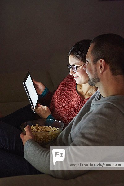 Mittelgroßes erwachsenes Paar sitzt abends auf dem Sofa  isst Popcorn und schaut auf ein digitales Tablet