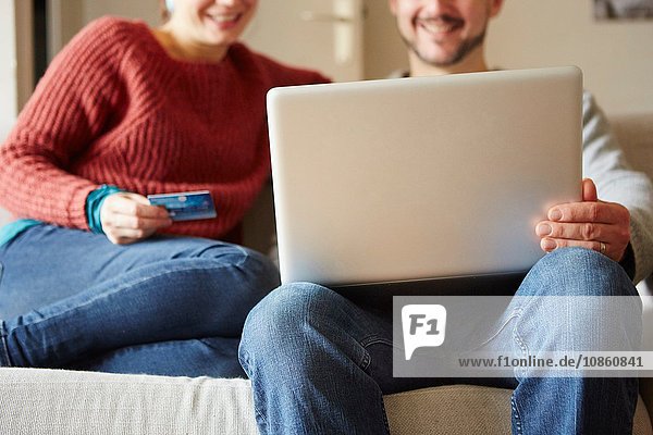 Cropped view of couple sitting on sofa using laptop and credit card