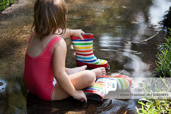 Kind in Gummistiefeln sitzt in Wasserpfütze