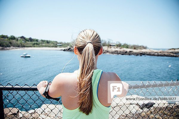 Rear view of woman wearing activity tracker holding wire fence looking at view of ocean