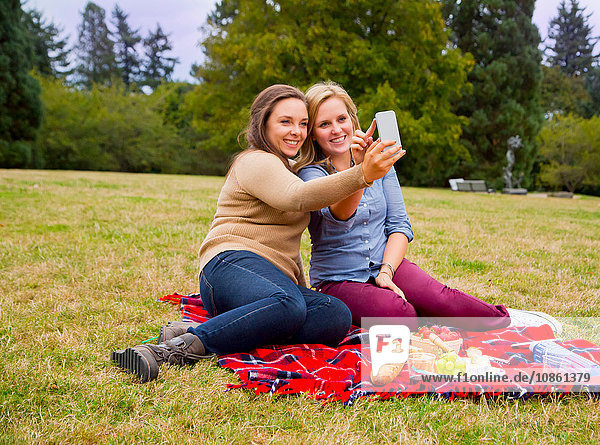 Two young women enjoying picnic in park  taking selfie  using smartphone