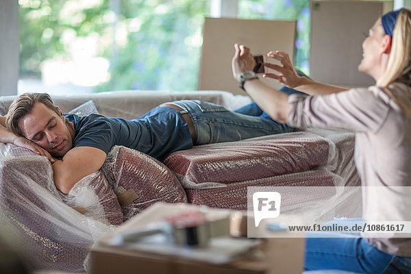 Umzug: Mann schläft auf einem mit Seifenblasen umhüllten Sofa  Frau fotografiert ihn  benutzt ein Smartphone